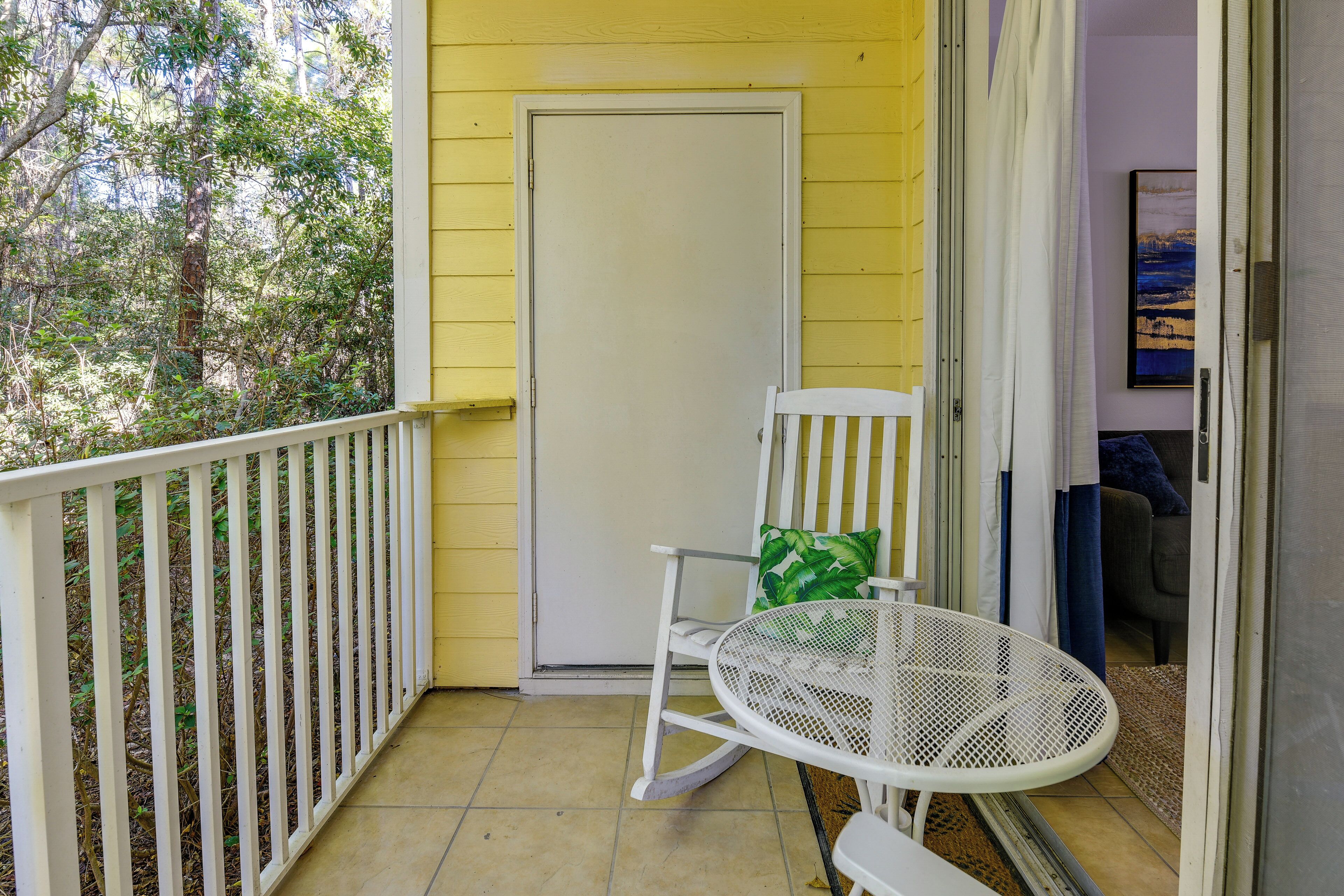 A balcony with a white rocking chair and a small table, a door, and a view of trees.