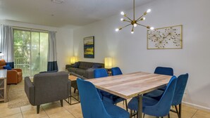 A modern living room with a dining area, featuring a wooden table, blue chairs, a sofa, and a ceiling light fixture. - Prime Miramar Beach Getaway w/ Pool Access! (Miramar Beach)