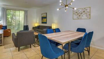 A modern living room with a dining area, featuring a wooden table, blue chairs, a sofa, and a ceiling light fixture.