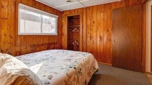 A bedroom with wooden walls, a bed with a floral bedspread, a window, and a closet with a door. - Lovely Au Gres Cottage w/ Hot Tub: Walk to Beach! (Au Gres)