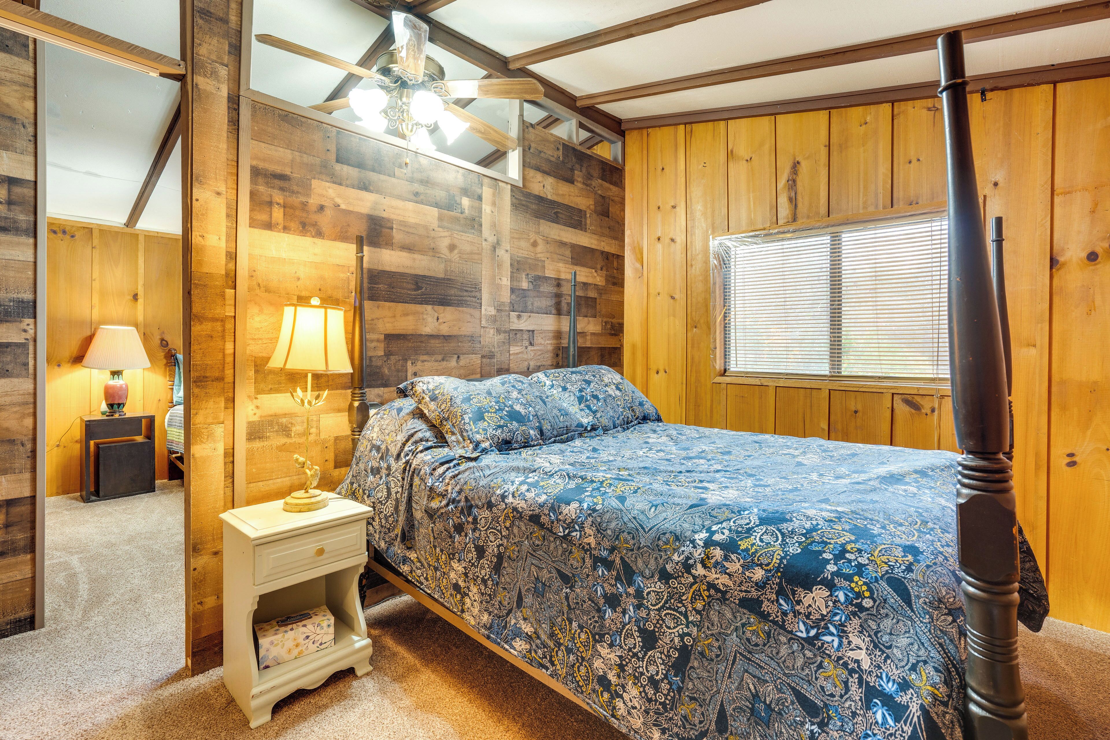 Un dormitorio con una pared de madera, una cama con colcha azul estampada, una mesita de noche con lámpara y un ventilador de techo.