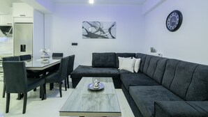 A modern living room with a sectional sofa, a dining table with chairs, a coffee table, a wall clock, and abstract art. - Jensila Suite (Zakynthos)