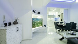 A modern kitchen with white cabinets, a dining area with a glass table, and a TV mounted on the wall displaying a landscape. - Jensila Suite (Zakynthos)