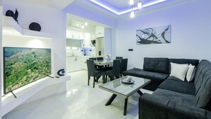 A modern living room with a glass coffee table, a dark grey sofa, and a flat-screen TV mounted on a white console. - Jensila Suite (Zakynthos)