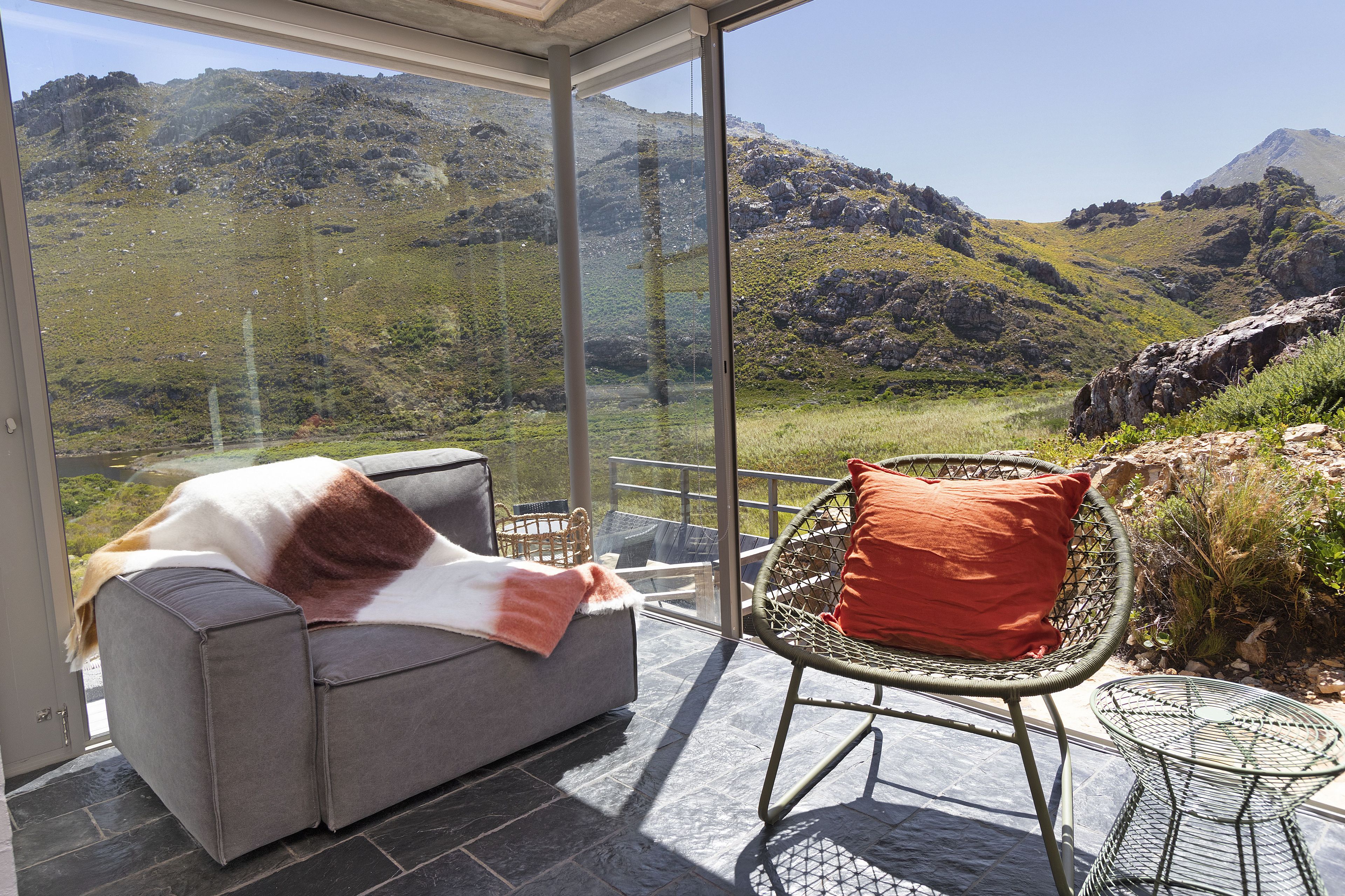 A modern room with a glass wall offering a mountain view, a grey sofa, and a wicker chair.