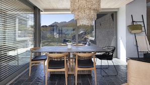 A modern dining area with a large table and chairs, a hanging light fixture, and a view of mountains through large windows. - Hytte Riverview (Rooiels)
