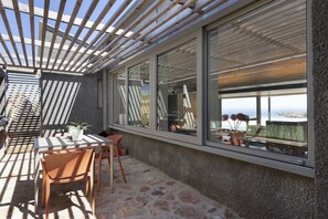 Un área de estar al aire libre con techo, una mesa y sillas, ventanas amplias y vista al mar.