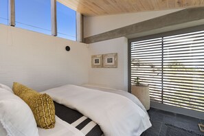 A bedroom with a large bed, a window with blinds, and a small wooden side table. - Hytte Riverview (Rooiels)