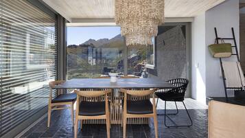 A modern dining area with a large table and chairs, a hanging light fixture, and a view of mountains through large windows.