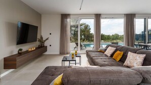 Un salón moderno con un televisor de pantalla plana grande, un sofá seccional gris con cojines amarillos, una pequeña mesa de centro y vistas a una piscina y follaje exterior.