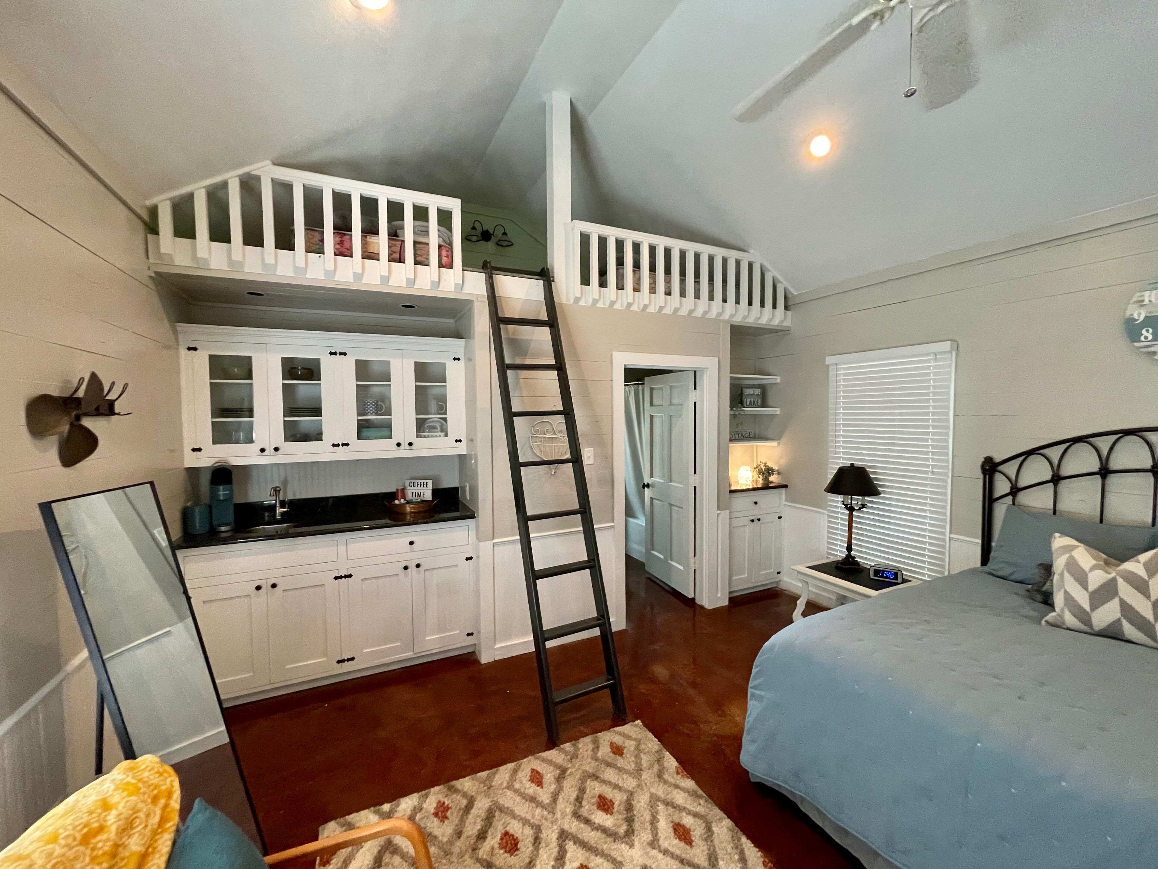 A bedroom with a lofted kitchen, a bed with patterned pillows, a nightstand with a lamp, and a wall clock.
