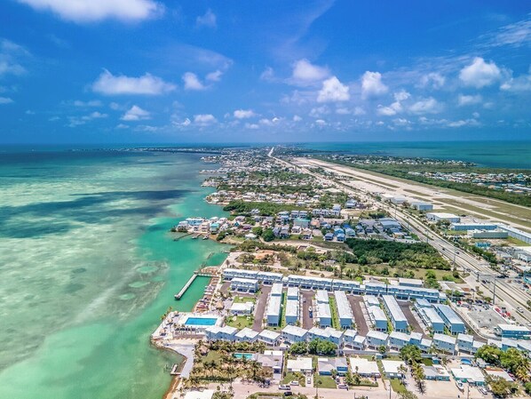 Vue aérienne d’une zone côtière comprenant un port de plaisance, un hôtel composé de plusieurs bâtiments et une route qui longe le littoral.