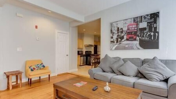 A living room with a grey sofa, a wooden coffee table, and a small yellow chair. There is a painting of a street scene on the wall. - NEW! Boston Condo Near MBTA, 3Mi to Fenway! (U1) (Boston)