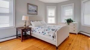A bedroom with a bed, bedside table, lamp, and a window with blinds. - NEW! Boston Condo Near MBTA, 3Mi to Fenway! (U1) (Boston)