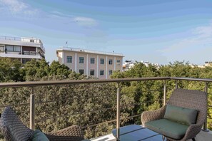 A balcony with wicker chairs and a table, overlooking a building and trees. - Phaedrus Living Heraklion City View Flat (Heraklion)