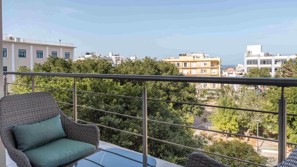 A balcony with wicker furniture and a glass table, overlooking a residential area with various buildings and trees. - Phaedrus Living Heraklion City View Flat (Heraklion)