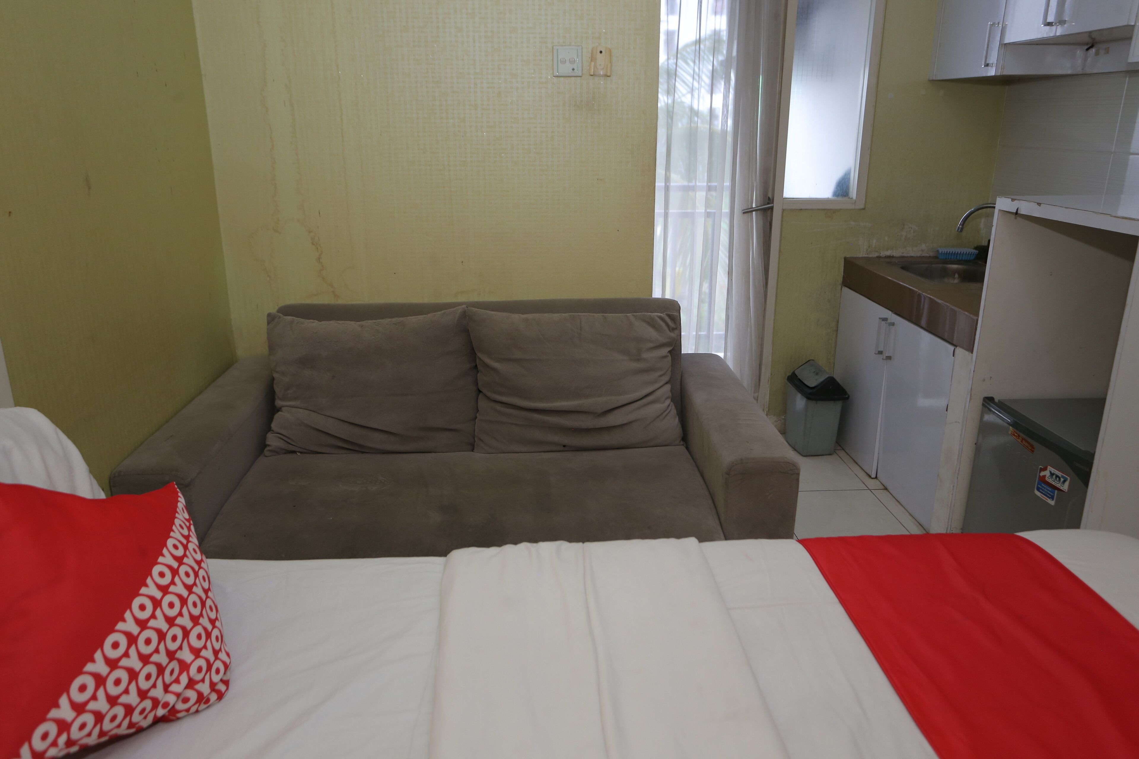 Quarto com cama, sofá, pequeno espaço de cozinha e janela com cortinas.