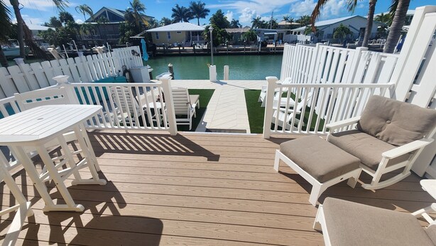 A wooden deck with white outdoor furniture, a view of a pool, and palm trees. - Paradise Found! Amazing 2/2 half duplex in KCB with Cabana Club & dockage (Key Colony Beach)