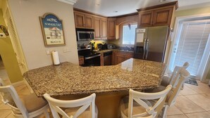 A kitchen with granite countertops, wooden cabinets, a stainless steel refrigerator, and a built-in microwave. There are two white chairs at the island. - Paradise Found! Amazing 2/2 half duplex in KCB with Cabana Club & dockage (Key Colony Beach)