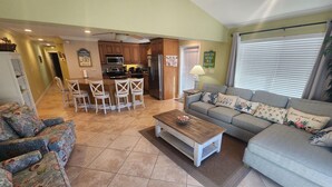A spacious living room with a grey sofa, a coffee table, and a patterned armchair. A kitchen with wooden cabinets and a dining area with white chairs is visible in the background. - Paradise Found! Amazing 2/2 half duplex in KCB with Cabana Club & dockage (Key Colony Beach)