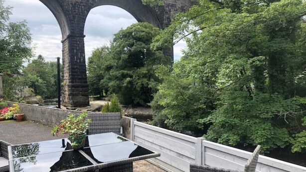 A patio with wicker furniture and a glass table, overlooking a stone bridge and lush greenery.