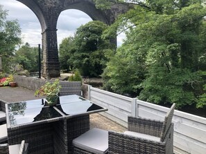 Een terras met rieten meubels en een glazen tafel, met uitzicht op een stenen brug en weelderige begroeiing.