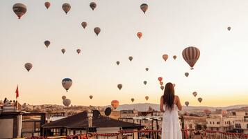 A rooftop terrace with a woman in a white dress overlooking a cityscape with numerous hot air balloons.