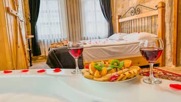 A hotel room with a bed, a table with fruit and wine glasses, and a stone wall.
