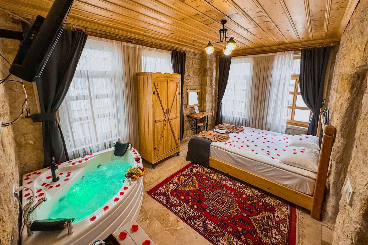 A room with a wooden ceiling, a bed with rose petals, a wooden wardrobe, a small table, and a bathtub with rose petals.