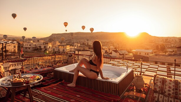 A woman sitting on a rooftop terrace with a hot tub, surrounded by a table with food and drinks, and hot air balloons in the background. - Tantan Cappadocia House (Nevsehir)