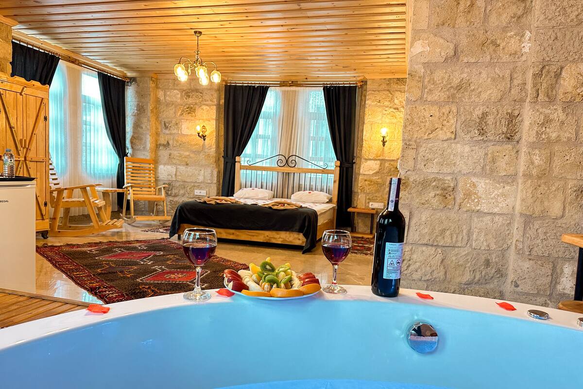 A bedroom with a large bed, a wooden ceiling, stone walls, and a hot tub with fruits and wine glasses.