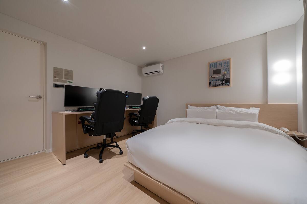 Habitación de hotel con cama, dos sillas de oficina negras, un escritorio con computadora, aire acondicionado y una mesita de noche con teléfono.