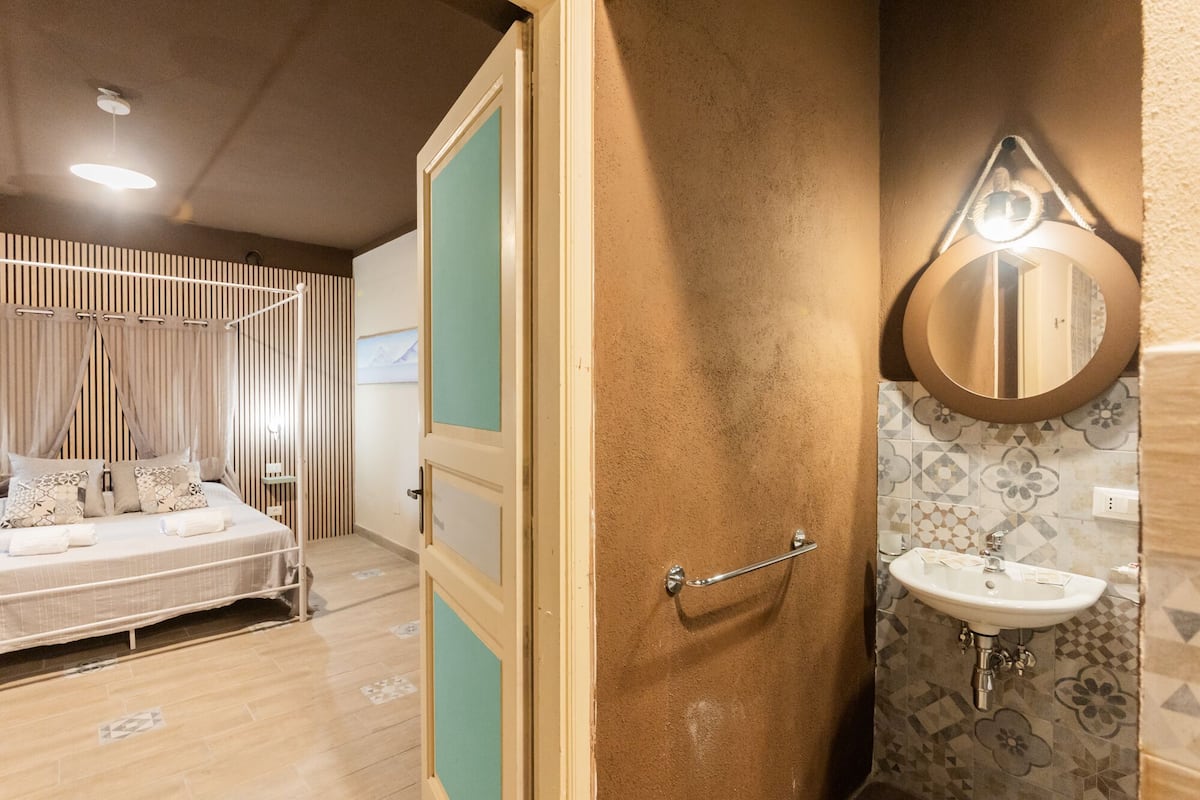 A bedroom with a bed, a bedside table, and a curtain. There is a bathroom with a sink and a mirror.