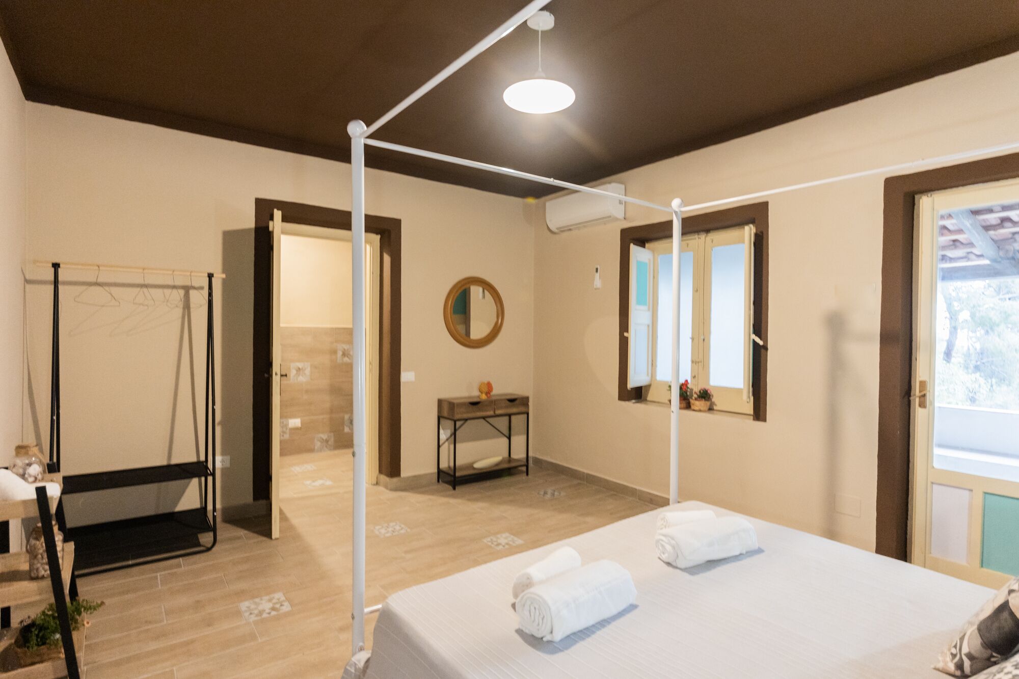 A hotel room with a bed, a bedside table, a mirror, a window, and a door leading to another room.