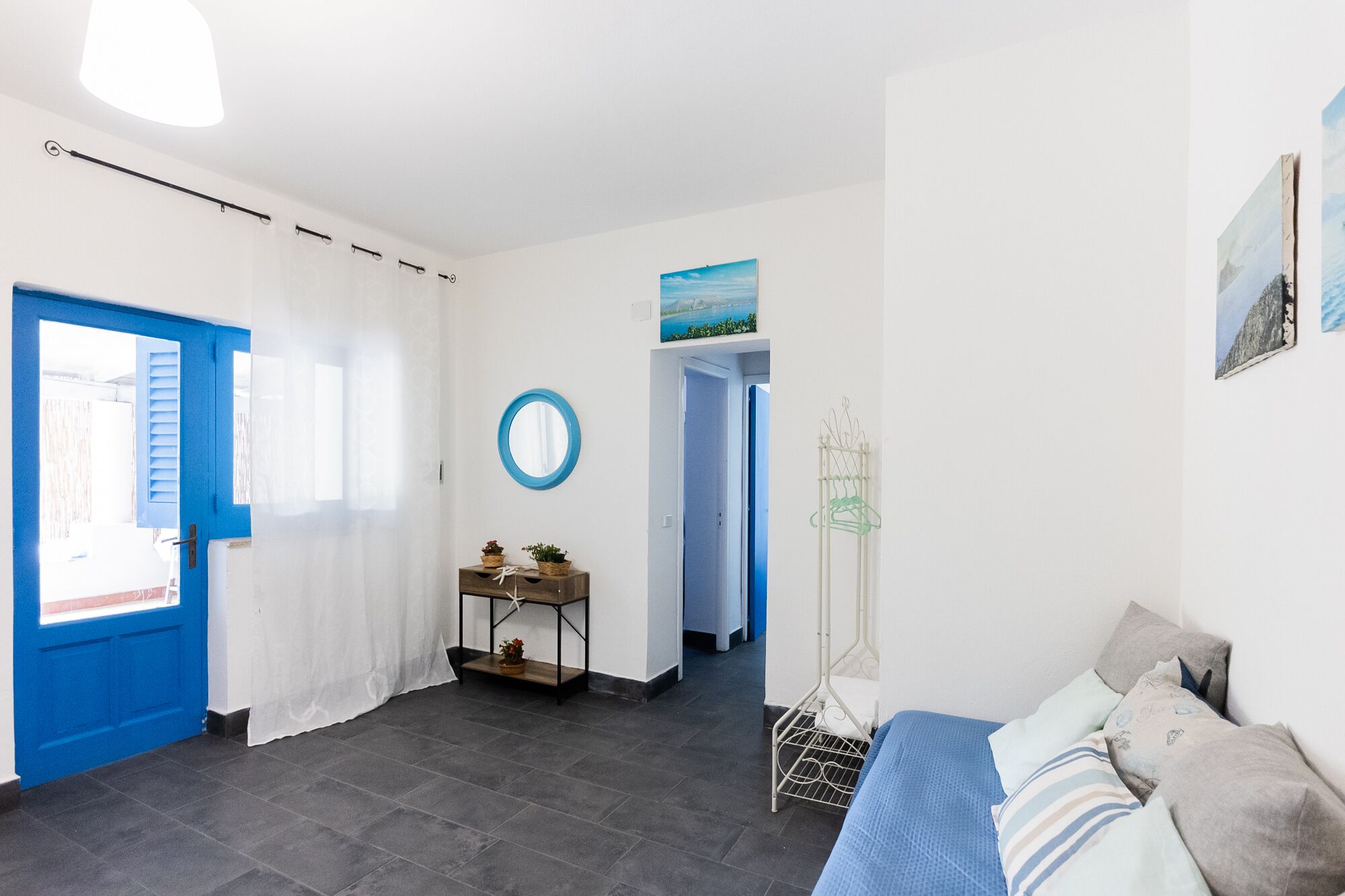 Una habitación con una puerta azul, un espejo redondo, una cama con almohadas, una mesita con plantas y vistas a un cielo azul a través de una ventana.