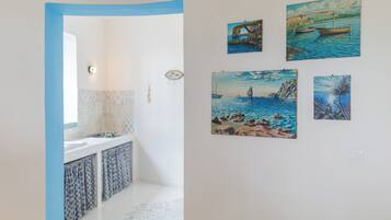 A bathroom with a white sink, a mirror, and a patterned shower area. There are four paintings on the wall depicting coastal scenes.