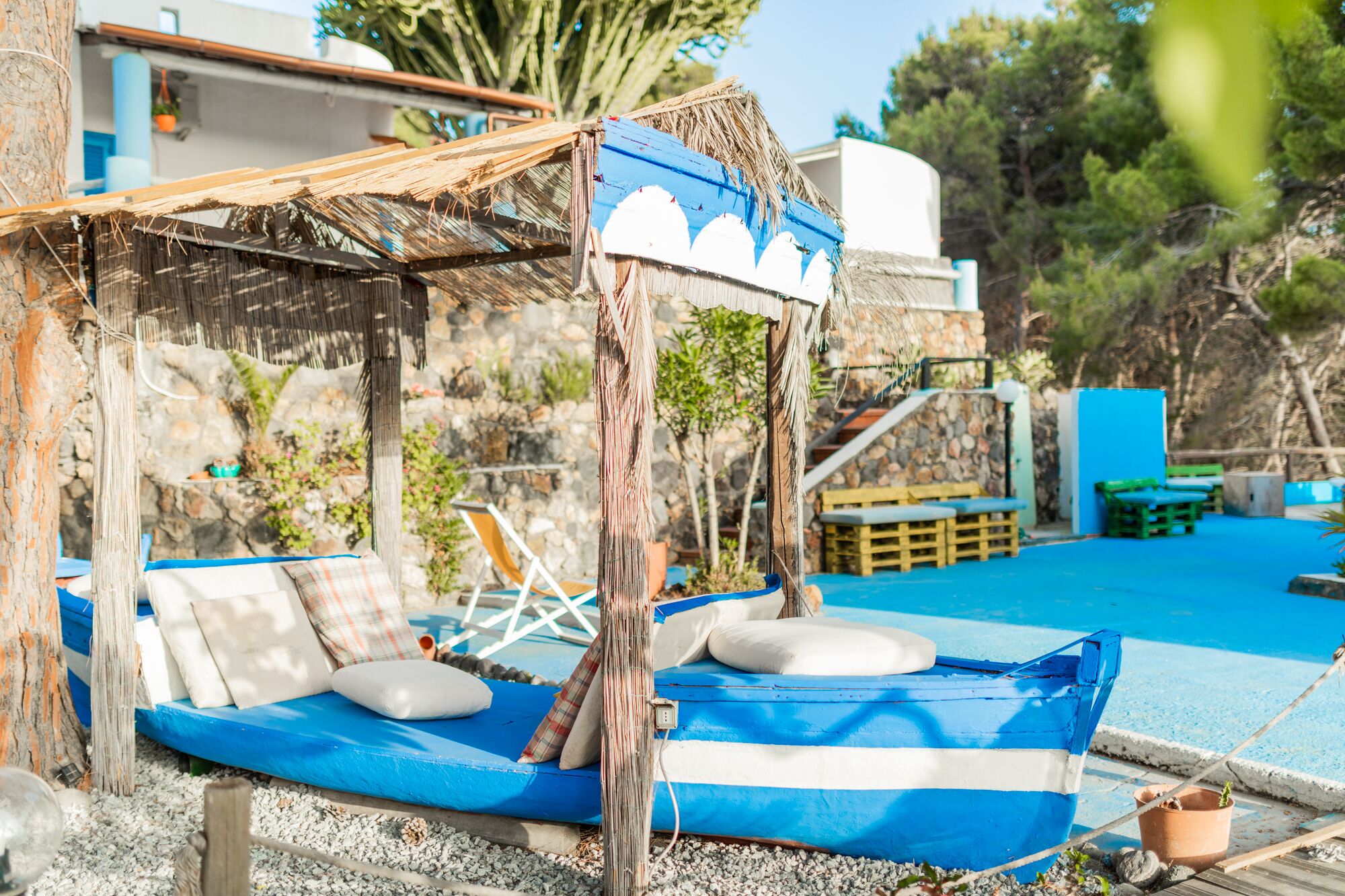 Un barco azul con cojines y una estructura de techo de paja, probablemente un área de estar, cerca de una piscina con un muro de piedra y follaje al fondo.