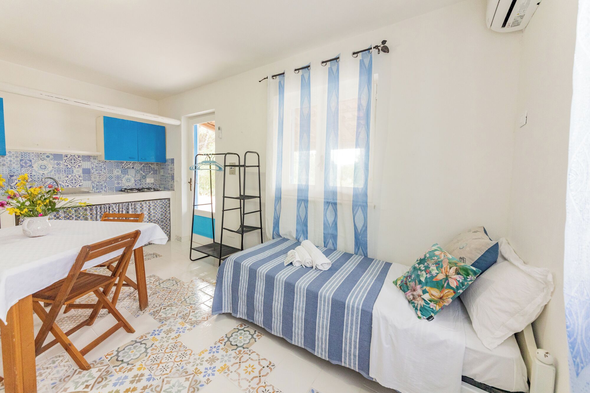 A compact room with a bed, a small table with chairs, a kitchen area with blue cabinets, and a window with blue curtains.