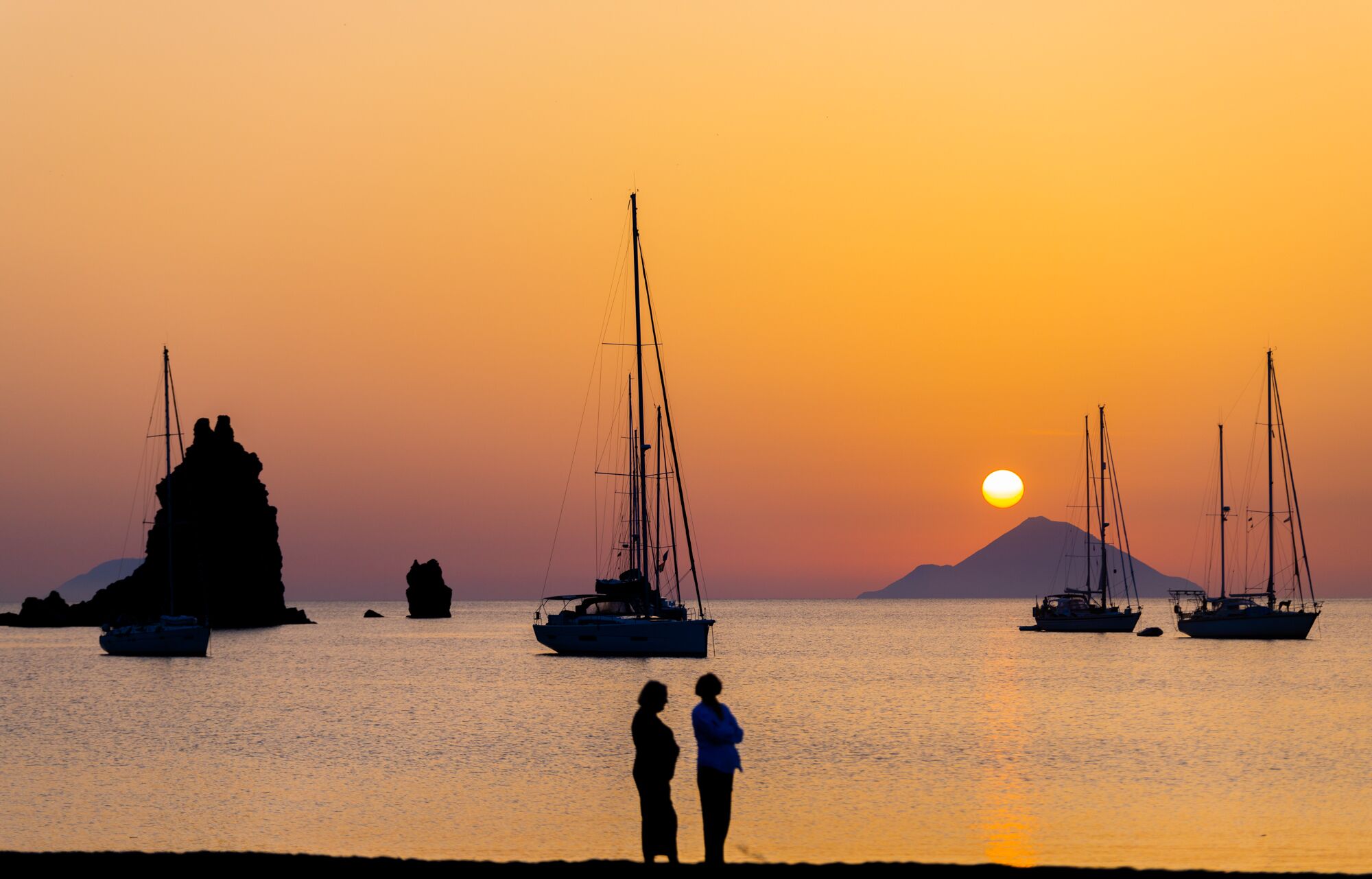 Un coucher de soleil sur une mer calme, avec plusieurs voiliers ancrés.