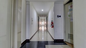 A long, narrow hallway with white walls and a black and white tiled floor. There are doors on both sides, a fire extinguisher mounted on the right wall, and a directional sign on the left wall. - OYO Life 92910 Apartemen Tamansari Panoramic By Tira (Bandung)