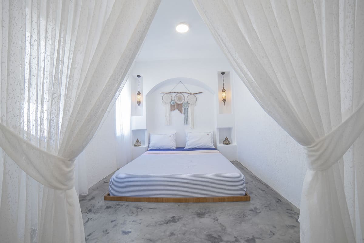 A bedroom with a bed, white curtains, wall sconces, and a decorative headboard.