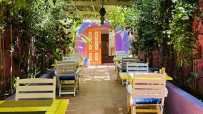 A covered outdoor seating area with wooden tables and chairs, surrounded by lush greenery and a colorful building in the background. - Casa De Nova Hotel (Bodrum)