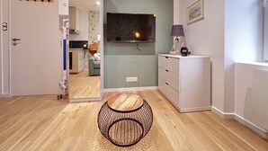 Un salon moderne avec un plancher en bois, une table ronde à structure métallique, un meuble blanc, un téléviseur à écran plat fixé au mur et une porte dotée d’un porte-manteau.