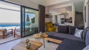 Una sala de estar moderna con un sofá, dos mesas de madera, una silla amarilla y vistas al mar a través de amplios ventanales.