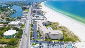 Vue aérienne d’une zone côtière avec une plage, une route et plusieurs bâtiments, dont un hôtel à la façade incurvée et un espace piscine.