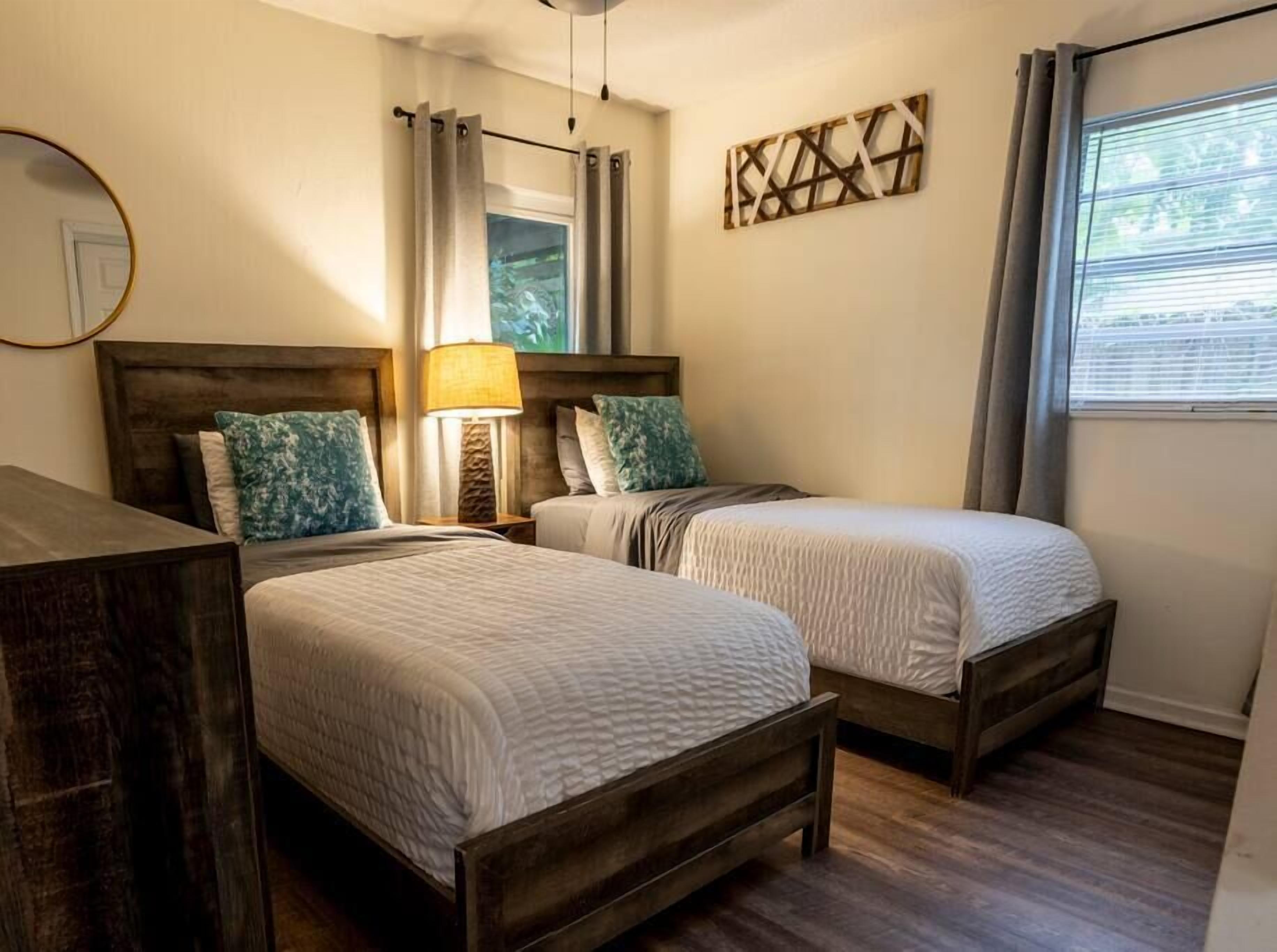 Een slaapkamer met twee bedden, een nachtkastje met een lamp, een spiegel en een raam met gordijnen.