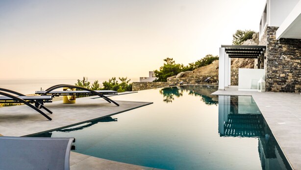 Un área de piscina moderna al aire libre con vista despejada al mar, un muro de piedra y mobiliario blanco.