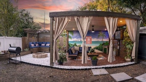 Un espacio al aire libre techado con un mural de una escena de playa, una parrilla y un conjunto de asientos con cojines y macetas.
