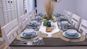 Una mesa de comedor dispuesta con platos azules y blancos, vasos transparentes y cubiertos, rodeada de sillas blancas.