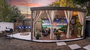 Un espace extérieur aménagé, abrité, avec une fresque représentant une scène de plage, un barbecue et un coin salon agrémenté de coussins et de jardinières.